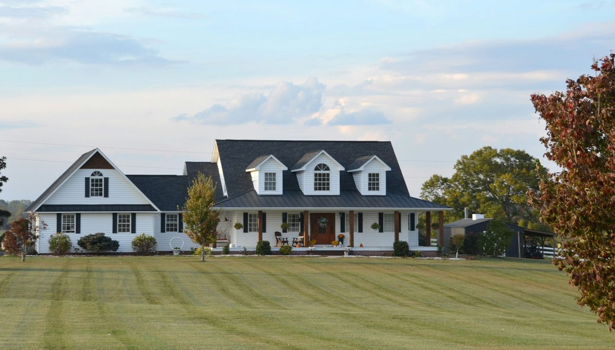 A white country farmhouse with dormer windows and a wrap-around front porch, set on a mowed green lawn in the golden light of early evening